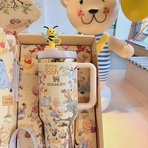 May include: A Stanley travel mug featuring Winnie the Pooh characters, with a yellow and black bee-shaped lid accent. The mug is in a decorative box with matching illustrations. A large bear statue is in the background.