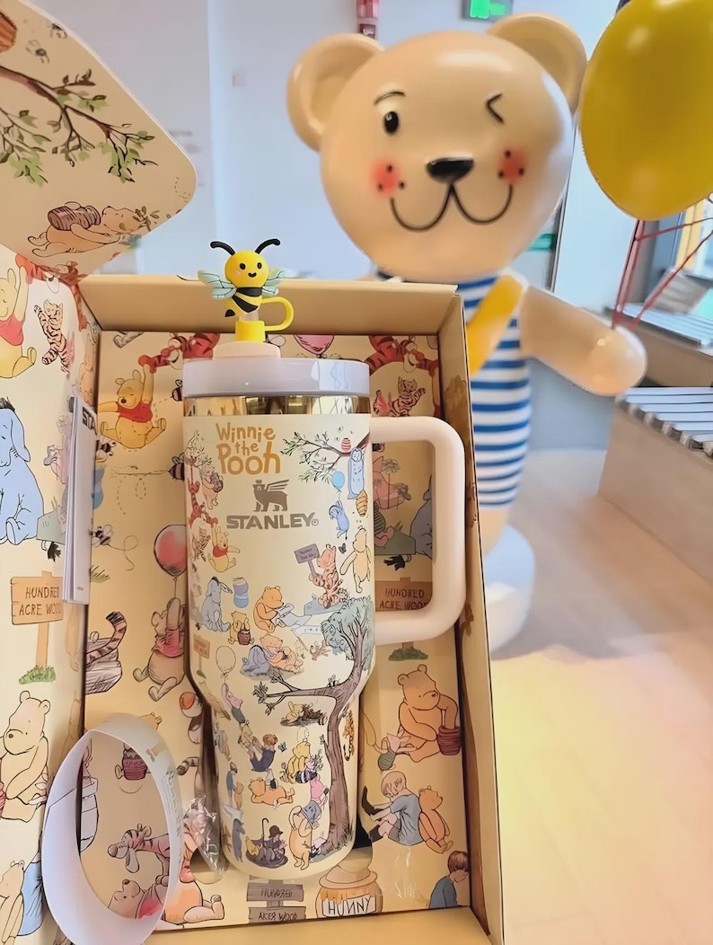 May include: A Stanley travel mug featuring Winnie the Pooh characters, with a yellow and black bee-shaped lid accent. The mug is in a decorative box with matching illustrations. A large bear statue is in the background.