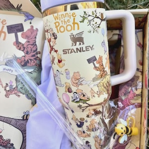 May include: A cream-colored Stanley tumbler featuring Winnie the Pooh characters and the text "Winnie the Pooh." The tumbler has a white handle and lid, with a gold band. The box includes a white shirt, a bee charm, and a straw.