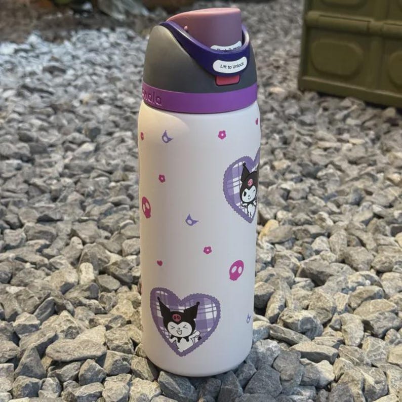 Puede incluir: Una botella de agua blanca con tapa morada, mecanismo de bot&oacute;n y pestillo de desbloqueo. La botella est&aacute; decorada con un patr&oacute;n repetitivo de peque&ntilde;as calaveras rosas, flores moradas y corazones grandes de cuadros morados con un personaje.
