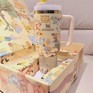 May include: A Stanley travel mug featuring Winnie the Pooh characters, with a yellow and white lid and a bee-shaped topper. The mug is in a matching box with illustrations of Pooh and friends.