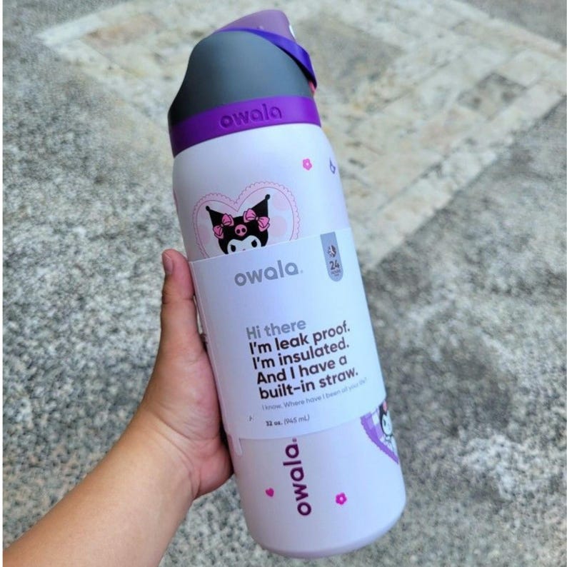 Puede incluir: Una botella de agua Owala de color morado claro con tapa gris oscuro y morada. La botella presenta un gr&aacute;fico del personaje Kuromi, peque&ntilde;as flores rosas y una etiqueta blanca que dice: Hi there, I'm leak proof. I'm insulated. And I have a built-in straw. 946 ml.