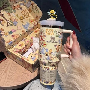 May include: A Stanley Winnie the Pooh themed travel mug with a white handle and a bee figurine on top. The mug is cream-colored and covered in illustrations of Winnie the Pooh characters. The mug is packaged in a matching box.
