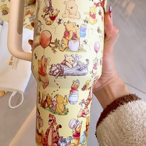 May include: Yellow tumbler featuring illustrations of Winnie the Pooh and friends. The tumbler has a cream-colored handle and the words "Winnie the Pooh" in a script font. The design includes characters like Pooh, Tigger, Piglet, and Eeyore.