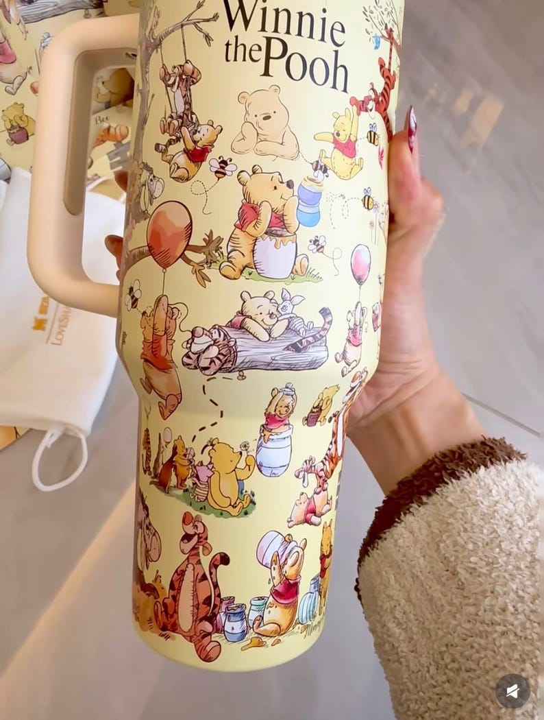 May include: Yellow tumbler featuring illustrations of Winnie the Pooh and friends. The tumbler has a cream-colored handle and the words "Winnie the Pooh" in a script font. The design includes characters like Pooh, Tigger, Piglet, and Eeyore.