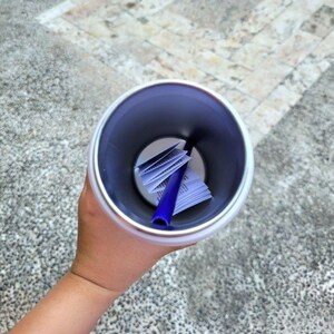 Puede incluir: Una vista desde arriba hacia el interior de un vaso cil&iacute;ndrico blanco con un interior met&aacute;lico liso. Dentro del recipiente, hay un documento de papel blanco plegado y una pajita de pl&aacute;stico azul s&oacute;lido apoyada en &aacute;ngulo contra la pared.