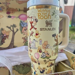 May include: A yellow Stanley tumbler featuring Winnie the Pooh characters. The tumbler has a white handle and lid, with the words "Winnie the Pooh" and the Stanley logo printed on the side. The tumbler is in a matching box.