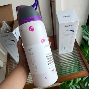 Puede incluir: Una botella de agua Owala FreeSip blanca con iconos de calaveras rosas, una tapa morada y gris, y un asa de transporte morada. La botella tiene una etiqueta con texto que incluye 'Hi there', 'I'm leak-proof', 'I'm insulated' y '32 oz. (945 ml)'.