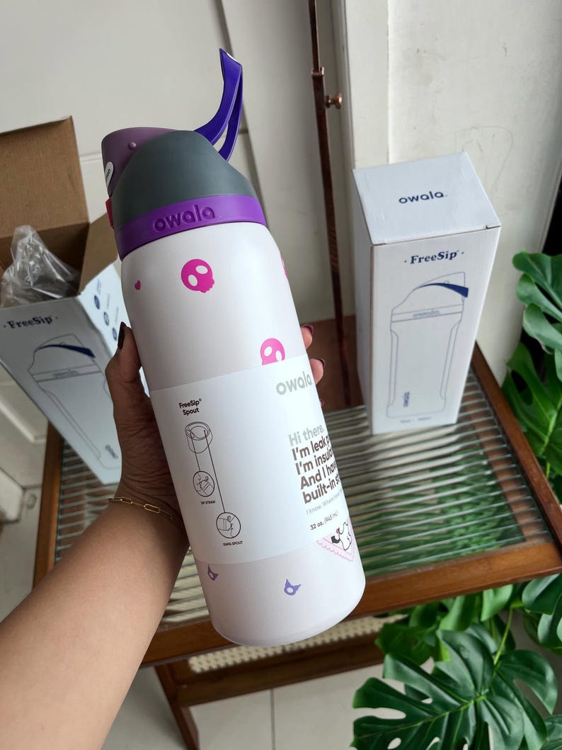 Puede incluir: Una botella de agua Owala FreeSip blanca con iconos de calaveras rosas, una tapa morada y gris, y un asa de transporte morada. La botella tiene una etiqueta con texto que incluye 'Hi there', 'I'm leak-proof', 'I'm insulated' y '32 oz. (945 ml)'.