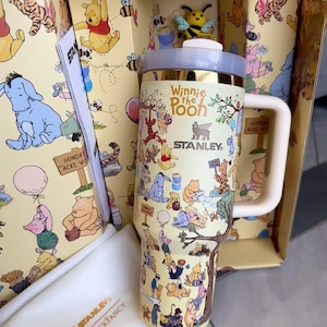 May include: A yellow Stanley travel mug featuring Winnie the Pooh characters. The mug has a white handle and a gold rim. The box and the mug are decorated with illustrations of Winnie the Pooh, Tigger, Piglet, and Eeyore. The text "Winnie the Pooh" and "Stanley" are visible.