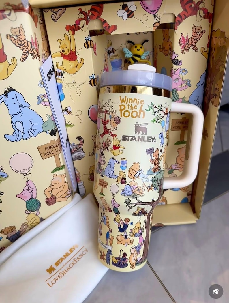 May include: A yellow Stanley travel mug featuring Winnie the Pooh characters. The mug has a white handle and a gold rim. The box and the mug are decorated with illustrations of Winnie the Pooh, Tigger, Piglet, and Eeyore. The text "Winnie the Pooh" and "Stanley" are visible.