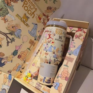 May include: A Winnie the Pooh themed Stanley tumbler and gift box. The tumbler is cream-colored with illustrations of Pooh and friends. The box features the same characters and the words "Winnie the Pooh" and "Stanley". A small bee figurine is included.