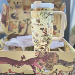 May include: A yellow Stanley travel mug featuring Winnie the Pooh characters and the text "Winnie the Pooh." The mug has a white handle and lid, and is packaged in a matching box with illustrations. The mug is approximately 7 inches tall.