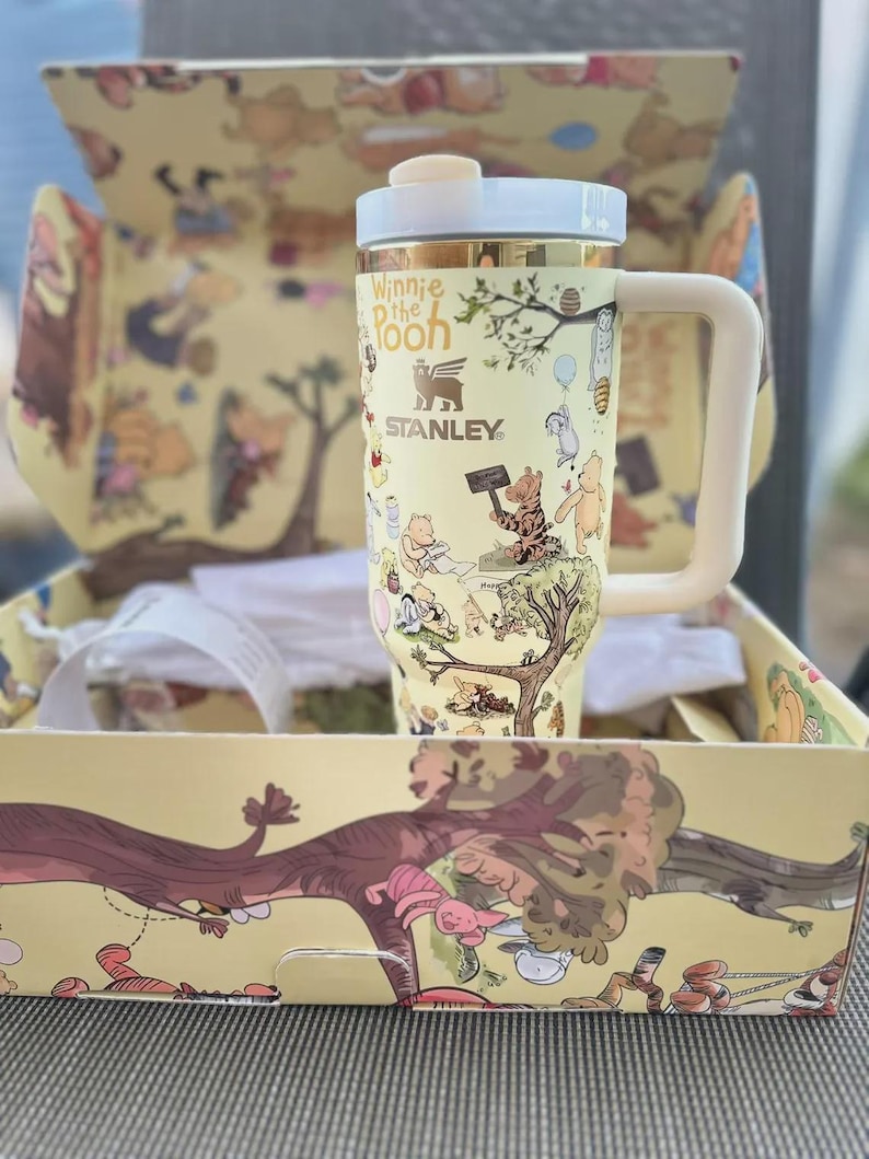 May include: A yellow Stanley travel mug featuring Winnie the Pooh characters and the text "Winnie the Pooh." The mug has a white handle and lid, and is packaged in a matching box with illustrations. The mug is approximately 7 inches tall.
