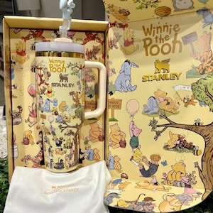 May include: A Winnie the Pooh themed Stanley tumbler with a white handle and lid, and a straw topper. The tumbler is in a yellow box with Winnie the Pooh illustrations. A white Loveshackfancy bag is in front.