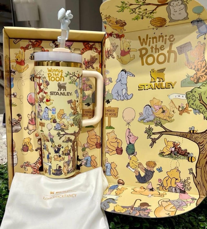 May include: A Winnie the Pooh themed Stanley tumbler with a white handle and lid, and a straw topper. The tumbler is in a yellow box with Winnie the Pooh illustrations. A white Loveshackfancy bag is in front.
