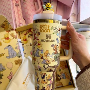 May include: A yellow Stanley tumbler featuring Winnie the Pooh characters. The tumbler has a white and gold lid, a cream-colored handle, and a bee-shaped topper. The design includes illustrations of Pooh, Piglet, Tigger, and Eeyore.