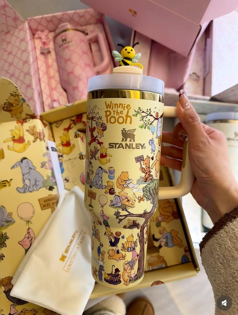 May include: A yellow Stanley tumbler featuring Winnie the Pooh characters. The tumbler has a white and gold lid, a cream-colored handle, and a bee-shaped topper. The design includes illustrations of Pooh, Piglet, Tigger, and Eeyore.