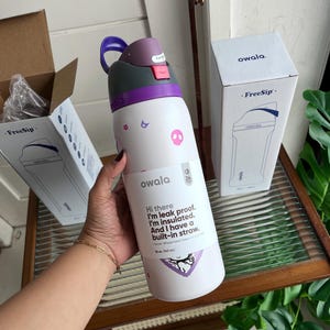 Puede incluir: Una botella de agua aislada Owala FreeSip blanca con tapa morada y rosa, decorada con peque&ntilde;os gr&aacute;ficos de calaveras y flores rosas. La etiqueta dice: Hi there I'm leak proof. I'm insulated. And I have a built-in straw. 32 oz. (945 mL).