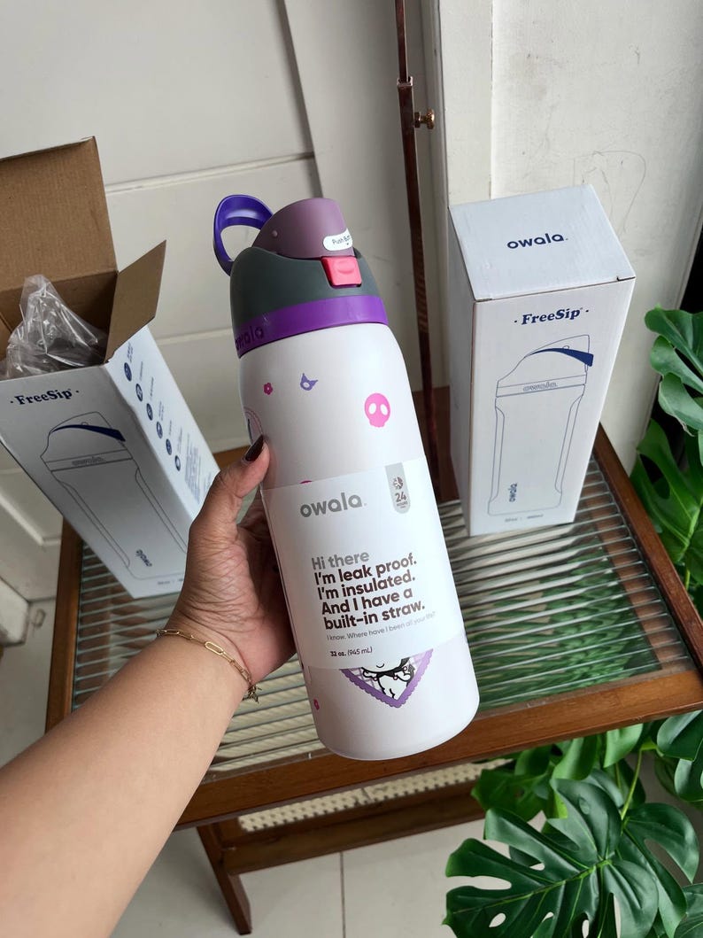 Puede incluir: Una botella de agua aislada Owala FreeSip blanca con tapa morada y rosa, decorada con peque&ntilde;os gr&aacute;ficos de calaveras y flores rosas. La etiqueta dice: Hi there I'm leak proof. I'm insulated. And I have a built-in straw. 32 oz. (945 mL).