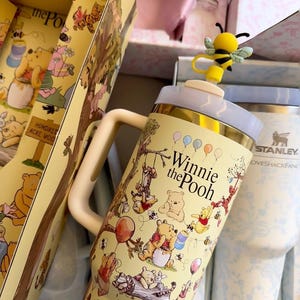 May include: A yellow Winnie the Pooh themed tumbler with a bee-shaped topper. The tumbler features illustrations of Pooh and friends, with the words "Winnie the Pooh" printed on the front. The tumbler is part of a gift set.