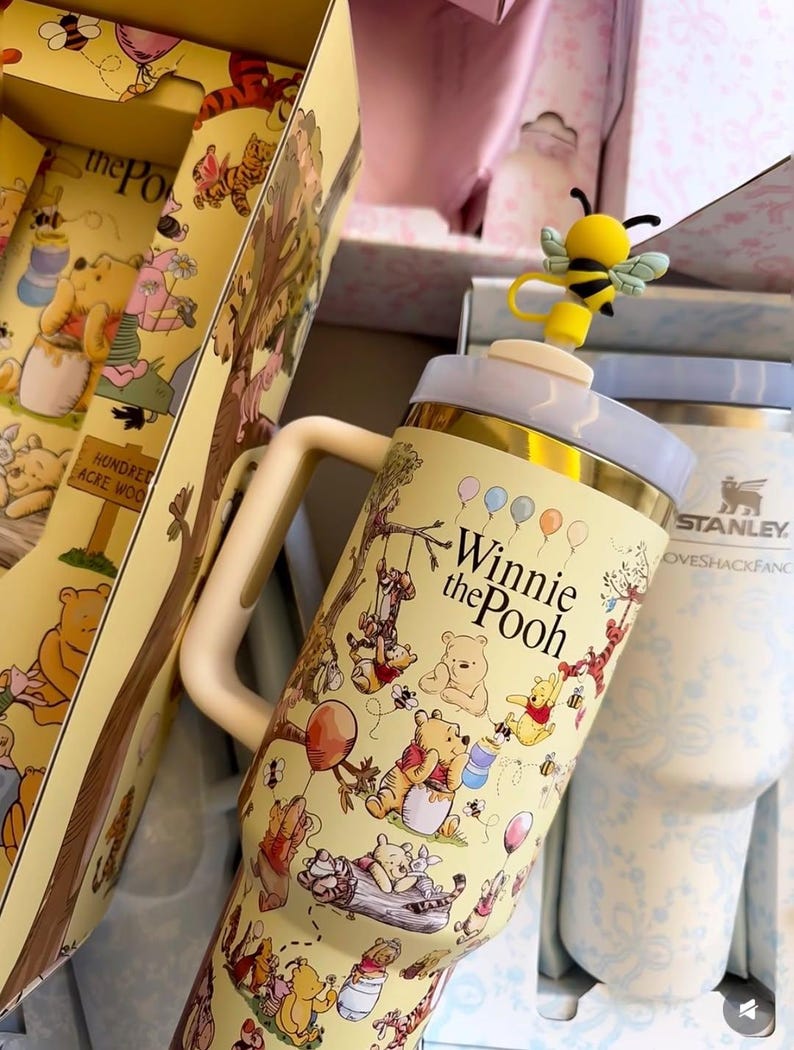 May include: A yellow Winnie the Pooh themed tumbler with a bee-shaped topper. The tumbler features illustrations of Pooh and friends, with the words "Winnie the Pooh" printed on the front. The tumbler is part of a gift set.