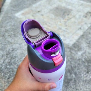 Puede incluir: Una botella de agua blanca con tapa morada y gris que incluye una tapa abatible y un cierre rosa. La tapa tiene una boquilla morada y una peque&ntilde;a pegatina con el texto Nicely done y Free Spirit. El cuerpo de la botella es blanco con iconos.