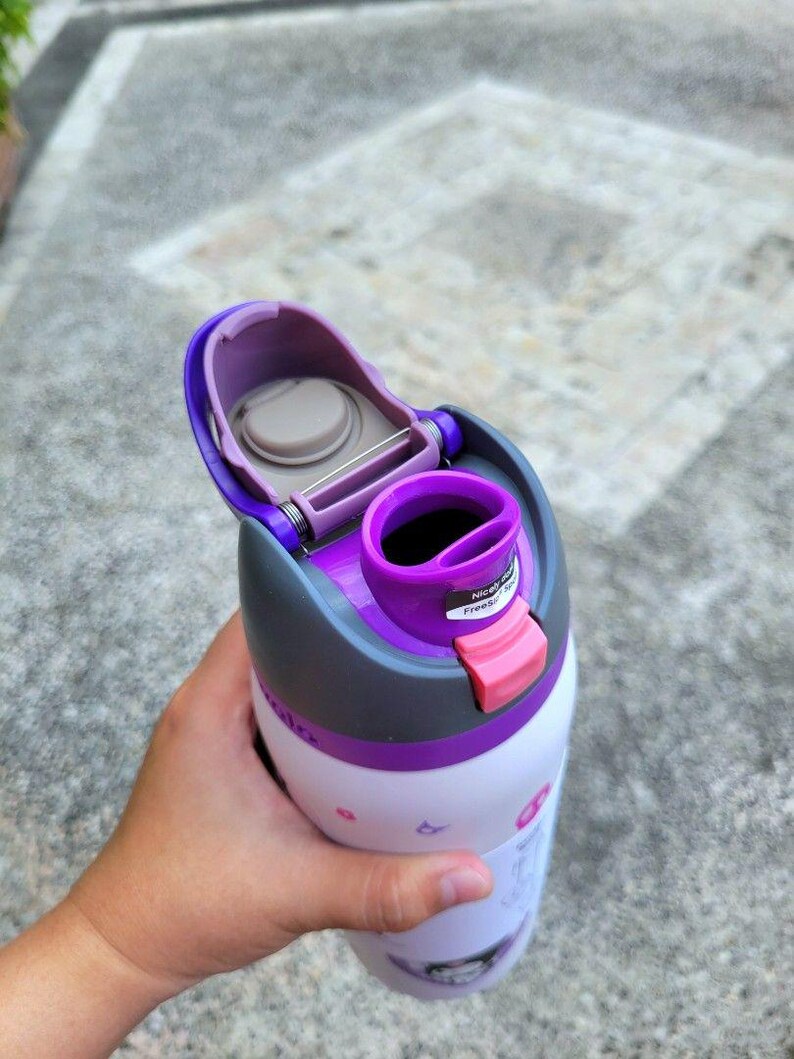 Puede incluir: Una botella de agua blanca con tapa morada y gris que incluye una tapa abatible y un cierre rosa. La tapa tiene una boquilla morada y una peque&ntilde;a pegatina con el texto Nicely done y Free Spirit. El cuerpo de la botella es blanco con iconos.