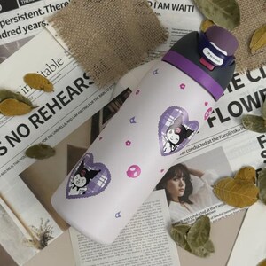 Puede incluir: Una botella de agua blanca con tapa morada y un mecanismo de apertura con la etiqueta "Lift to Unlock". La botella presenta un patr&oacute;n repetitivo de peque&ntilde;as flores rosas, calaveras rosas y dos pegatinas moradas en forma de coraz&oacute;n.