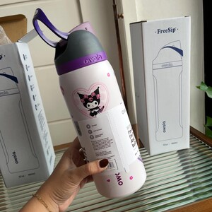 Puede incluir: Una botella de agua Owala FreeSip blanca con asa de transporte morada y tapa gris. La botella presenta un gr&aacute;fico del personaje Kuromi dentro de un coraz&oacute;n rosa, peque&ntilde;os iconos de flores rosas y una etiqueta informativa blanca alrededor.