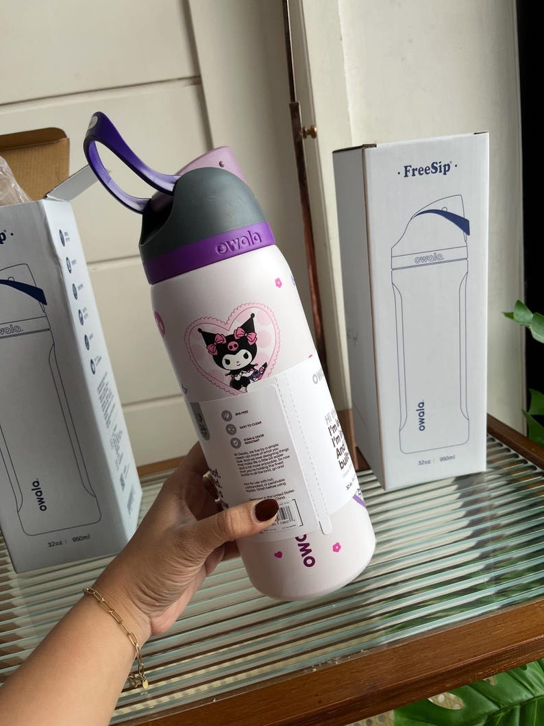 Puede incluir: Una botella de agua Owala FreeSip blanca con asa de transporte morada y tapa gris. La botella presenta un gr&aacute;fico del personaje Kuromi dentro de un coraz&oacute;n rosa, peque&ntilde;os iconos de flores rosas y una etiqueta informativa blanca alrededor.