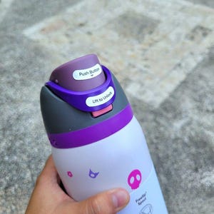 Puede incluir: Una botella de agua blanca con tapa morada y gris que incluye un bot&oacute;n y un mecanismo de desbloqueo. La botella tiene peque&ntilde;os iconos gr&aacute;ficos rosas y morados, incluyendo una calavera, y el texto FreeSip Spout con un diagrama.