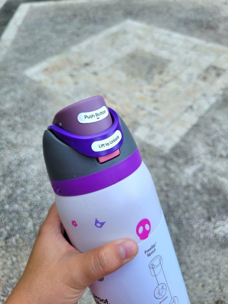 Puede incluir: Una botella de agua blanca con tapa morada y gris que incluye un bot&oacute;n y un mecanismo de desbloqueo. La botella tiene peque&ntilde;os iconos gr&aacute;ficos rosas y morados, incluyendo una calavera, y el texto FreeSip Spout con un diagrama.