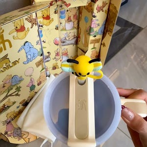 May include: A Winnie the Pooh themed gift set featuring a light blue and cream-colored bowl with a bee-shaped handle. The set includes a box with illustrations of Winnie the Pooh characters, a cream-colored bag, and other accessories.
