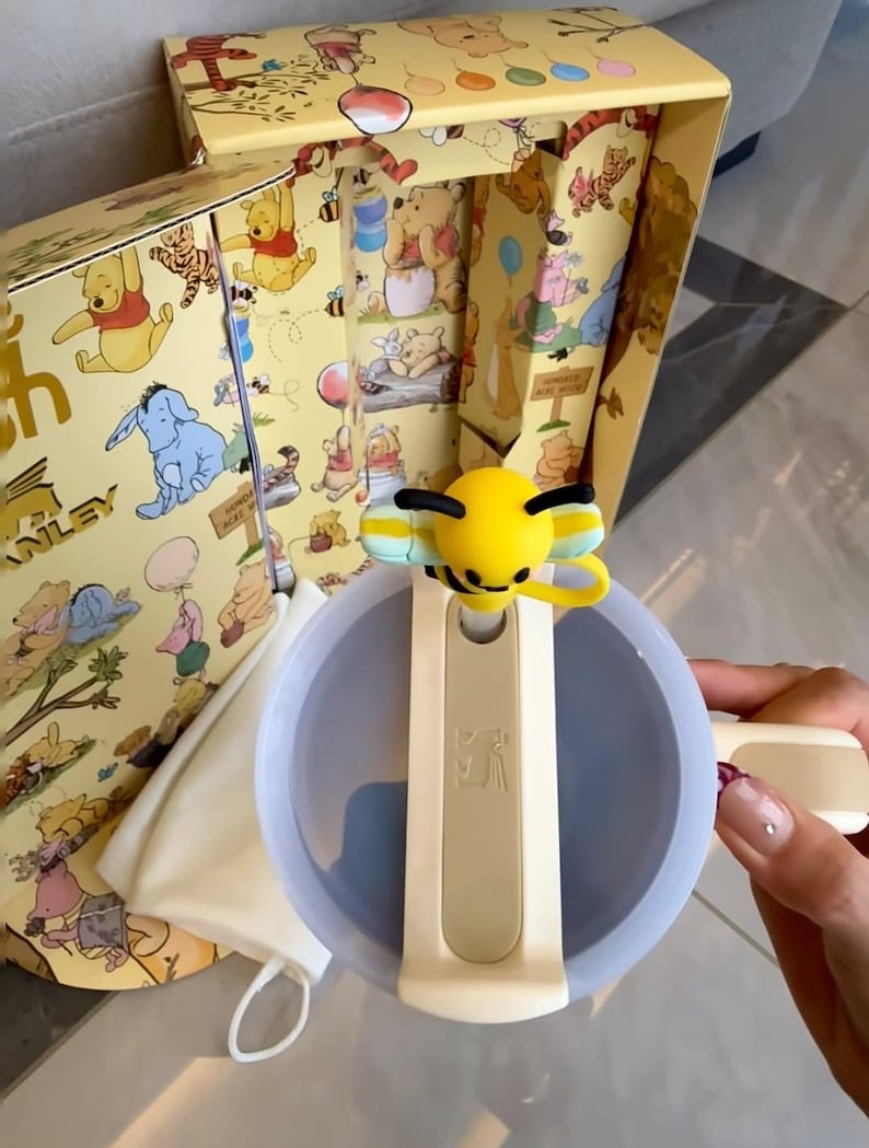 May include: A Winnie the Pooh themed gift set featuring a light blue and cream-colored bowl with a bee-shaped handle. The set includes a box with illustrations of Winnie the Pooh characters, a cream-colored bag, and other accessories.