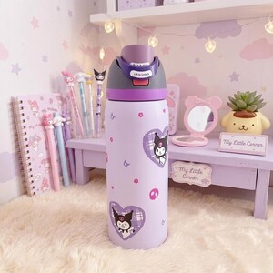 Puede incluir: Una botella de agua de color morado claro con un gr&aacute;fico del personaje Kuromi, patrones en forma de coraz&oacute;n y peque&ntilde;os iconos de calaveras rosas. La tapa es de color morado oscuro y gris con un bot&oacute;n que dice Push Button y Lift to Unlock.