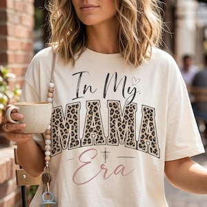 Puede incluir: Una camiseta de manga corta color crema con el texto negro In My y un pequeño corazón, seguido de la palabra MAMA en una fuente grande con estampado de leopardo, y la palabra Era en cursiva rosa con un pequeño símbolo de cruz encima.