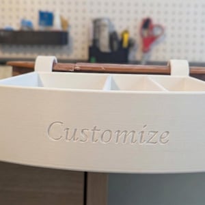 Può includere: Un organizer rettangolare bianco stampato in 3D, dotato di tre scomparti interni e due ganci per appenderlo. La parte anteriore presenta la parola Customize in rilievo con un carattere serif. La finitura è liscia e opaca.