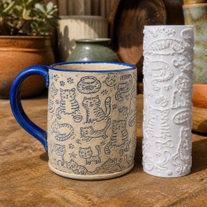 May include: A ceramic mug with a beige base and blue cat-themed illustrations, including fish bowls and paw prints, featuring a solid blue handle. Beside it stands a white cylindrical rolling pin embossed with a repeating pattern of cats, fish, and the word Kitty.