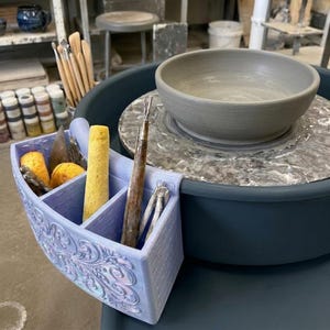 May include: A light purple, 3D-printed pottery tool organiser with an ornate, embossed scrollwork pattern on the front. The organiser is attached to a dark blue pottery wheel, holding a yellow sponge and metal sculpting tools near a clay bowl.