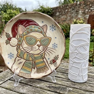 Op de afbeelding: Een rond keramieken bord met een beschilderde kat die een rode kerstmuts, een groen mutsje met patroon, een zonnebril met geel montuur en een gestreepte sjaal draagt, naast een wit cilindervormig 3D-geprint object met een reliëf van een kat.