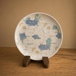 May include: A round white ceramic plate featuring a dense, embossed pattern of various stylized cats in shades of blue, green, and yellow. The cats are depicted with diverse facial expressions and postures, creating a textured, layered surface design.