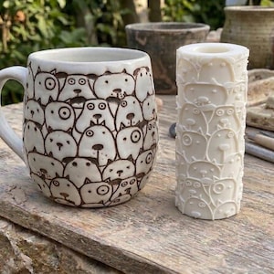 May include: A white ceramic mug and a matching cylindrical clay texture roller, both featuring a repeating pattern of hand-drawn style dog faces with dark outlines. The mug has a curved handle, while the roller is a solid white tool.