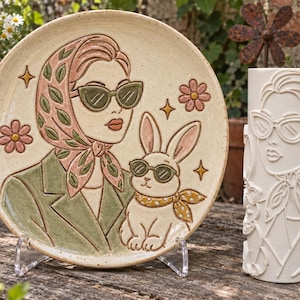 Può includere: Un piatto rotondo in ceramica con un rilievo colorato di una persona con foulard e occhiali da sole accanto a un coniglio, accompagnato da un vaso cilindrico in ceramica bianca abbinato con un design in rilievo monocromatico identico.