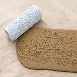 May include: A white cylindrical clay rolling pin with embossed text and symbols, positioned next to a flat piece of tan clay that has been imprinted with the same pattern, including words like Faith, Hope, God, Grace, Prayer, and Blessed.