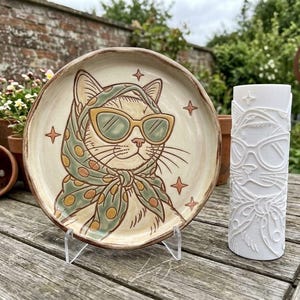 May include: A round ceramic plate featuring a hand-painted illustration of a cat wearing a green polka-dot headscarf and yellow-rimmed sunglasses, accompanied by a white cylindrical candle embossed with the same cat design and star accents.