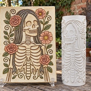 Può includere: Una placca rettangolare in ceramica con un rilievo di uno scheletro con capelli lunghi circondato da fiori rosa e foglie verdi, accanto a un vaso cilindrico in ceramica bianca con un identico motivo a rilievo di scheletro e fiori.
