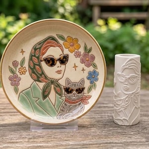 Può includere: Un piatto rotondo in ceramica con una colorata illustrazione in rilievo di una persona che indossa una giacca verde, un foulard fantasia e occhiali da sole leopardati, accompagnata da un gatto con occhiali simili, accanto a un vaso cilindrico bianco.