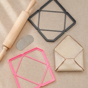Peut inclure: Un rouleau à pâtisserie en bois, une petite raclette ovale en métal, un emporte-pièce gris foncé en forme d'enveloppe, un emporte-pièce rose en forme d'enveloppe et une enveloppe en tissu beige pliée sur une surface texturée claire.