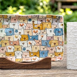 Könnte beinhalten: Ein rechteckiges Keramiktablett mit einem bunten, handbemalten Muster aus verschiedenen Hundegesichtern in Blau-, Gelb-, Rosa- und Weißtönen, präsentiert auf einem dunklen Holzständer neben einem weißen zylindrischen Keramik-Nudelholz mit Hundemotiven.